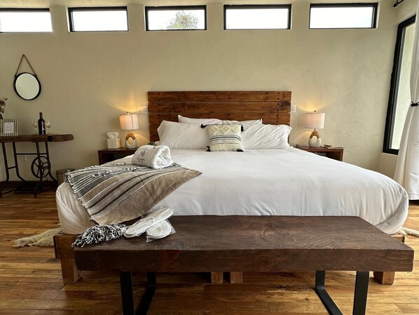 A neatly arranged bedroom with a large bed, wooden headboard, bedside tables with lamps, a small table with a bottle and glasses, and a bench.