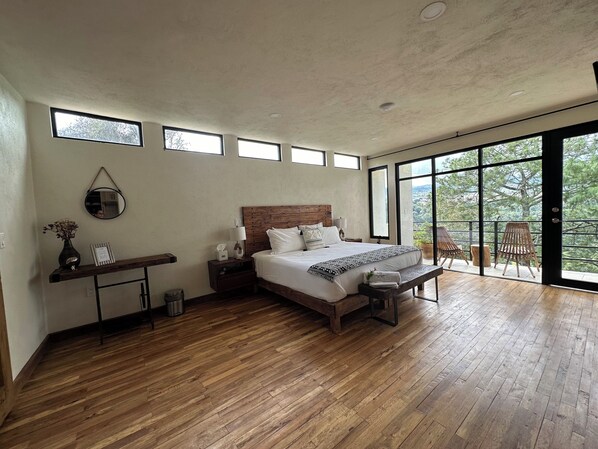 Un dormitorio amplio con una cama grande, suelo de madera, una mesita de noche con lámpara, un estante pequeño con objetos decorativos y vistas al exterior a través de ventanas grandes.