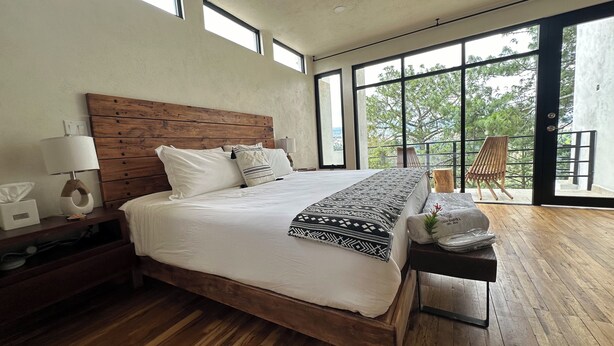 Een moderne slaapkamer met een groot bed, een houten hoofdbord en uitzicht op bomen door grote ramen.