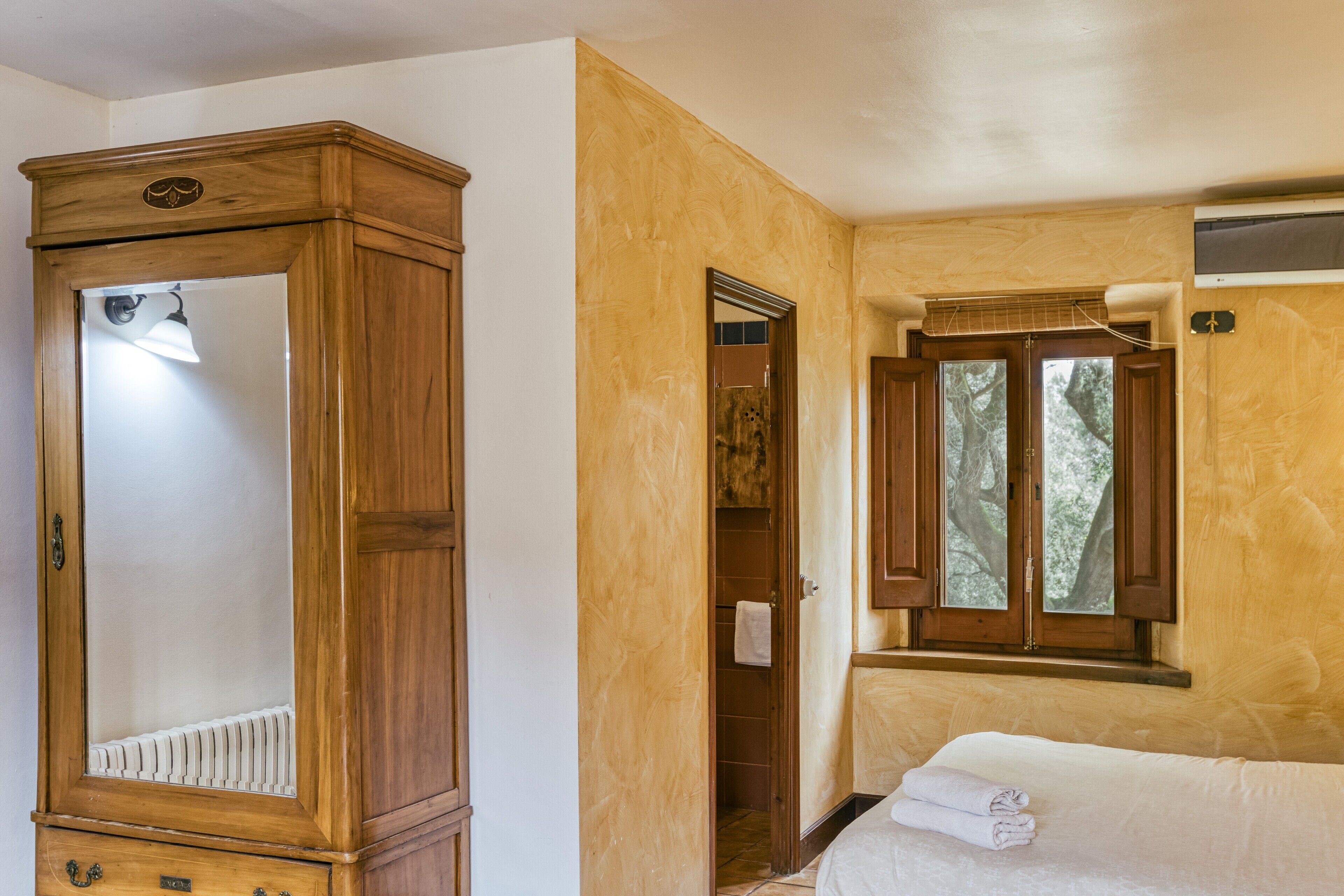 A room with a wooden wardrobe, a bed with a white mattress, a window with a view of trees, and a radiator under the wardrobe.