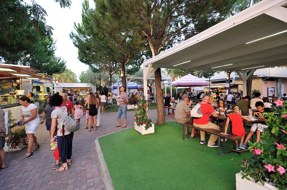 An outdoor market with people walking and sitting at tables, surrounded by trees and covered areas.