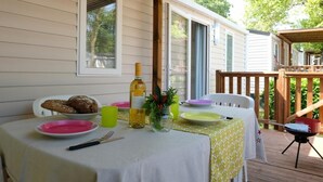 Une table à manger installée à l’extérieur, avec des assiettes, des couverts et une bouteille de vin, entourée d’une terrasse en bois et d’un espace abrité.