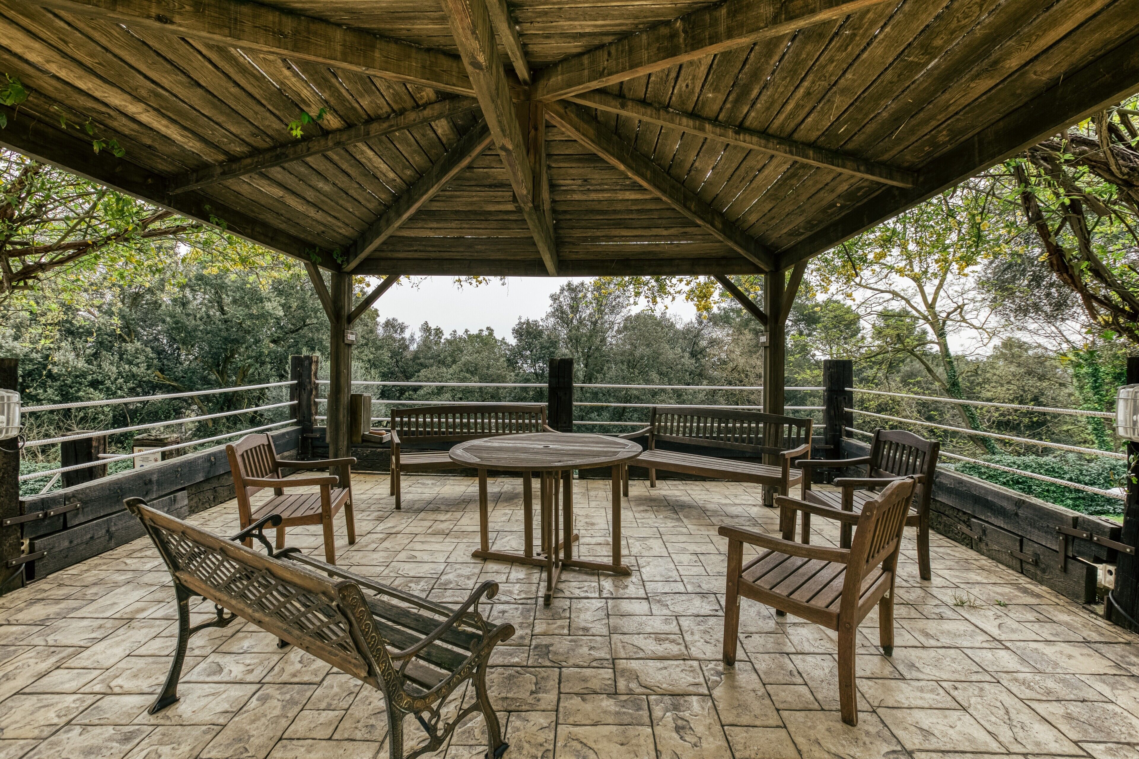 A covered outdoor seating area with stone flooring, wooden benches, and a central table.