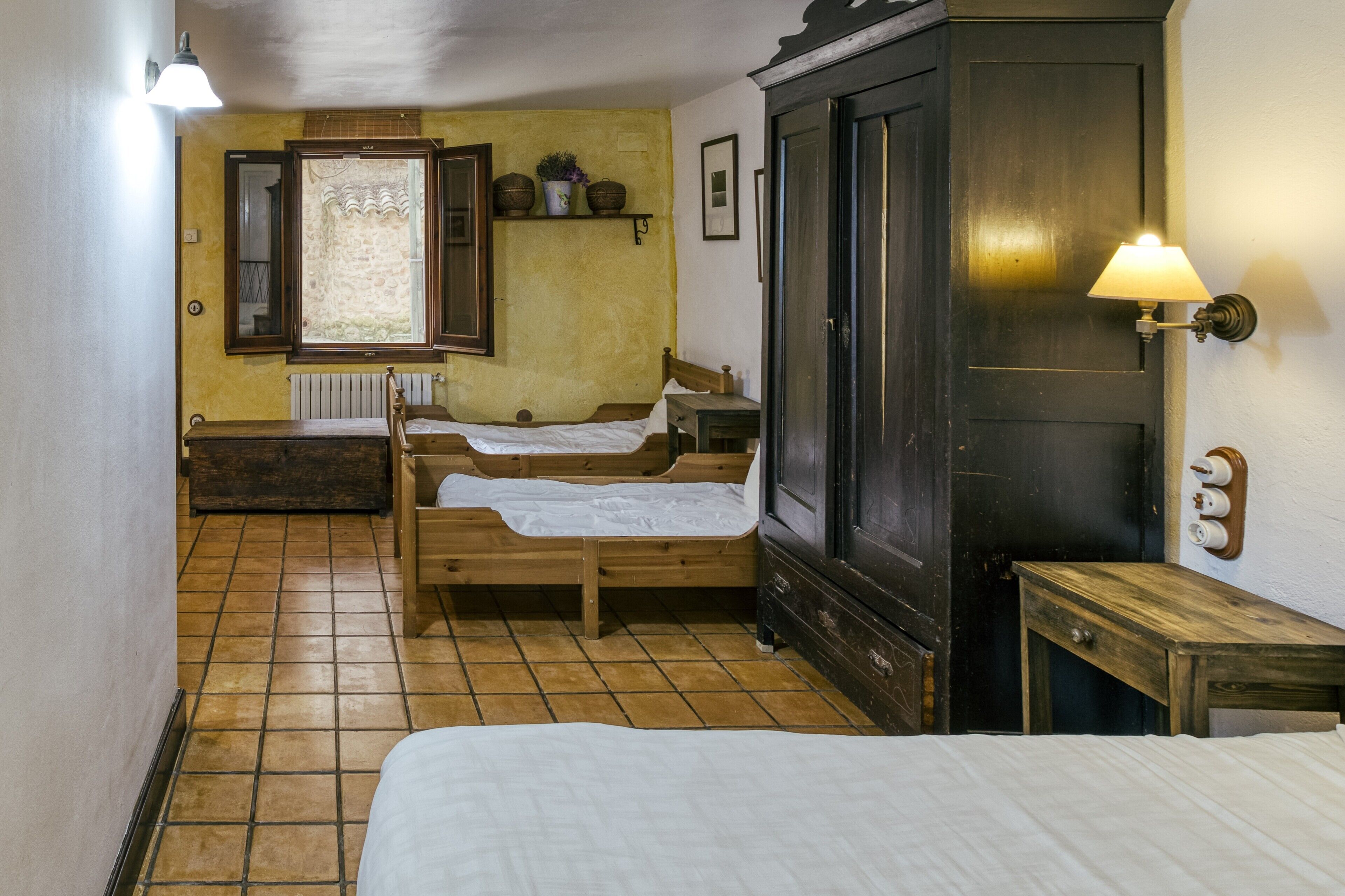 A room with two wooden beds, a dark wardrobe, a small table, and a wall-mounted shelf with baskets.