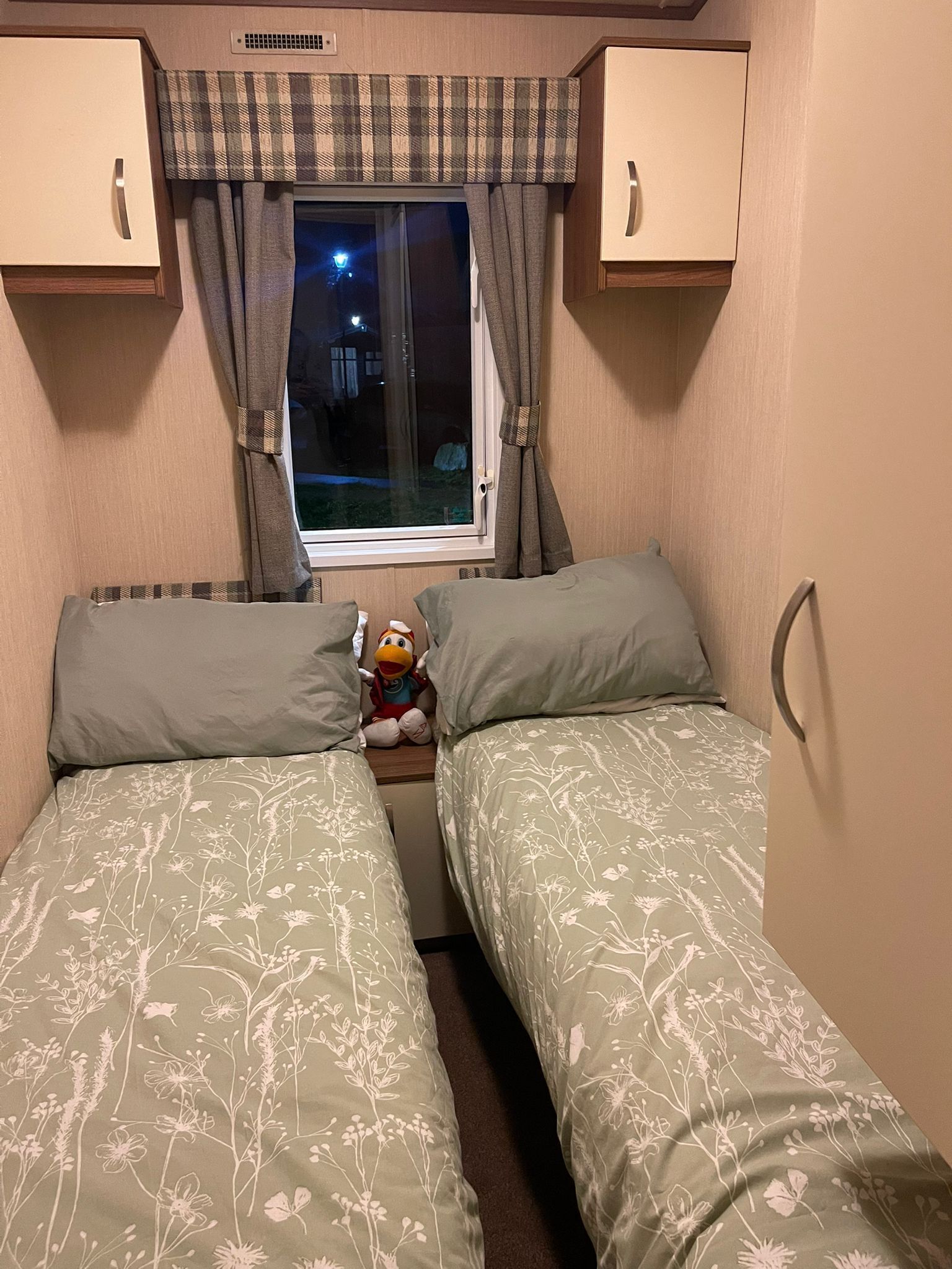 A small, cozy bedroom with two beds, a window with curtains, and a plush toy on the nightstand.