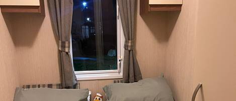 A small, cozy bedroom with two beds, a window with curtains, and a plush toy on the nightstand.