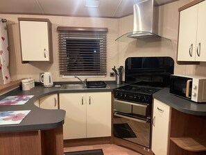 A compact kitchen with a stove, microwave, and sink. - 3 Bedroom Caravan (Grove 12) on Trecco Bay Holiday Park, Porthcawl (Porthcawl)