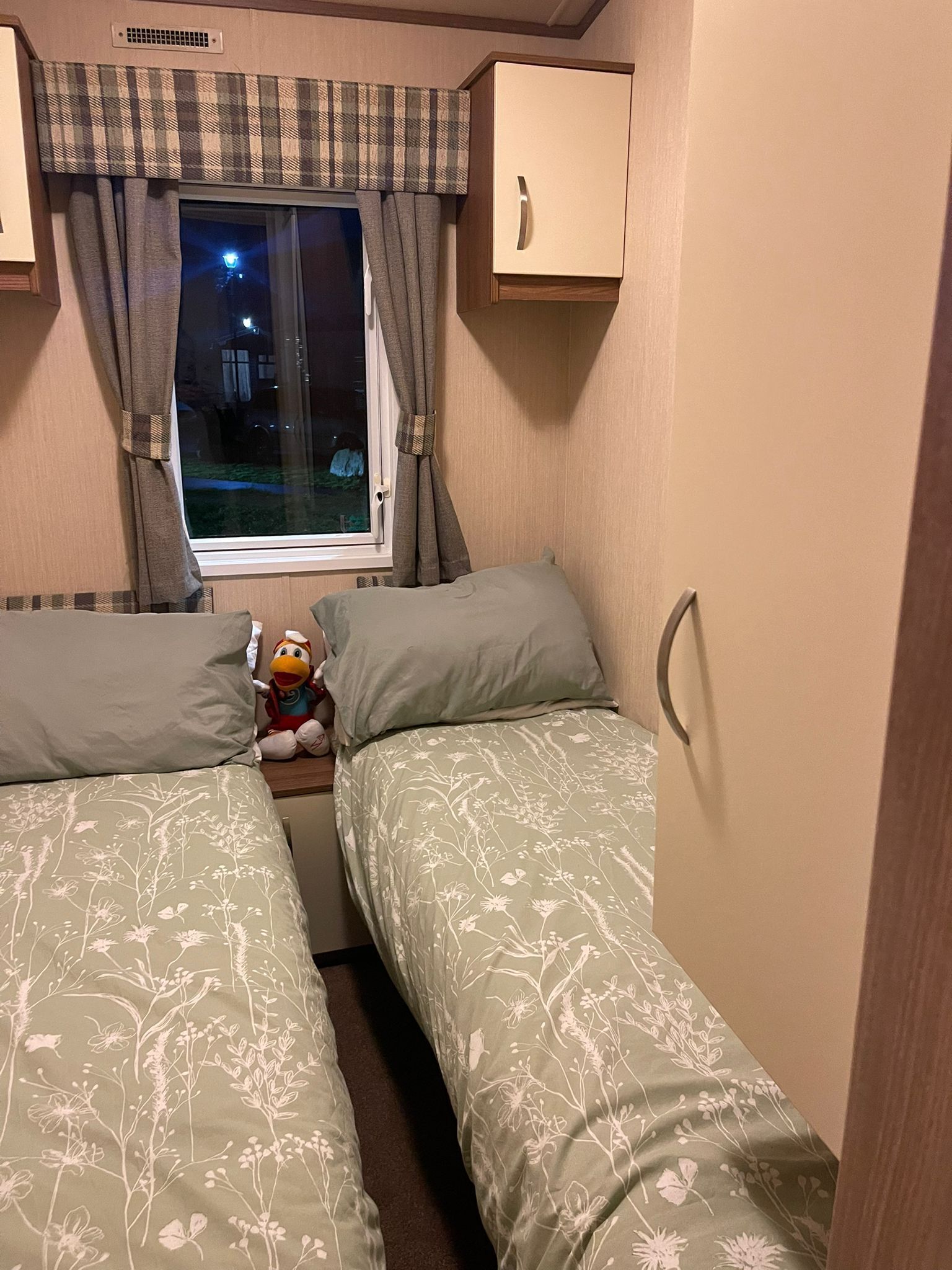 A small, cozy room with two single beds, a window with curtains, a plush toy on one bed, and a cabinet above the window.