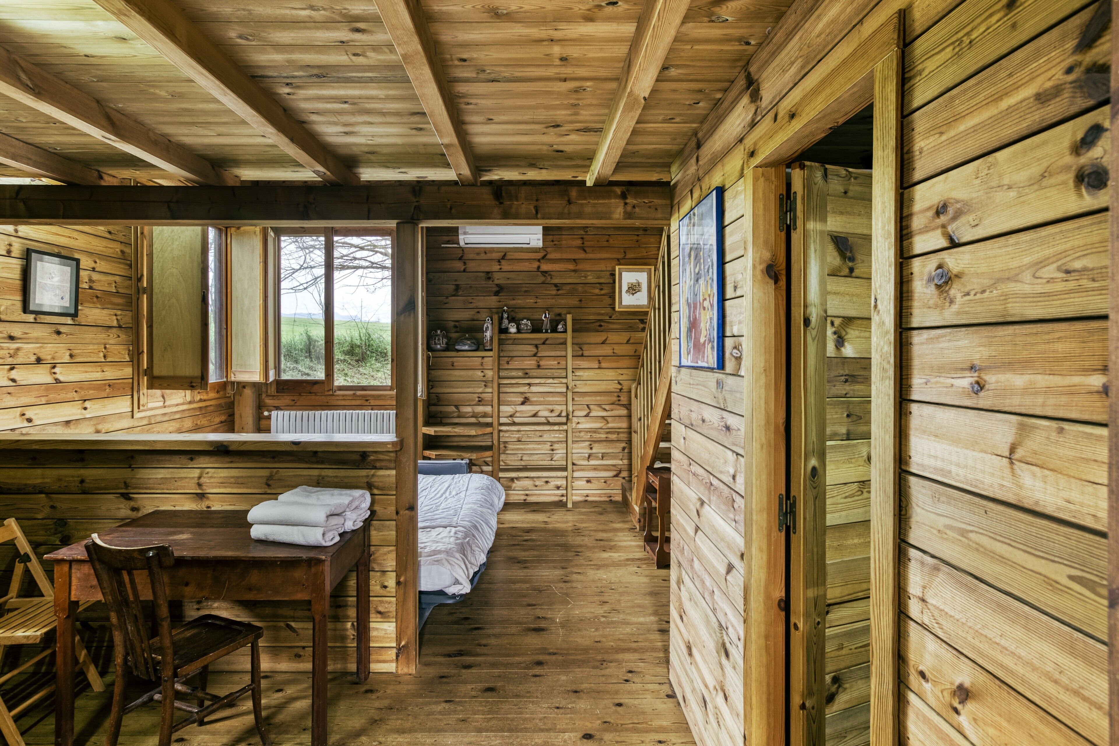 Interior de una cabaña de madera con una cama, una mesa y toallas, y una ventana.