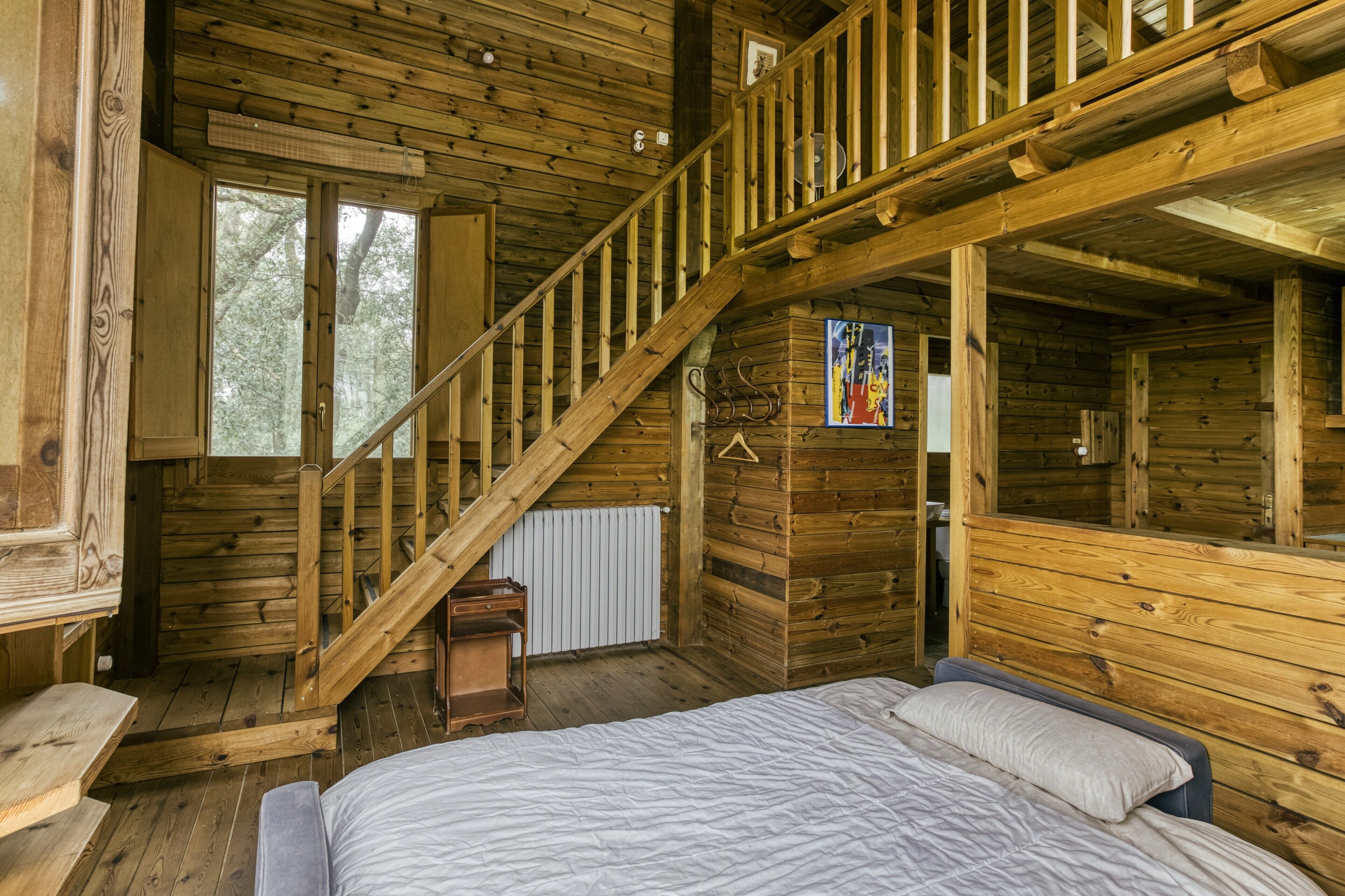 Interior de una cabaña de madera con una cama, una escalera y una ventana.