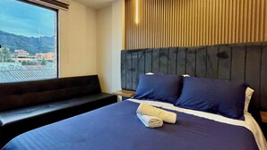 Une chambre d’hôtel moderne avec un grand lit, une tête de lit ornée de lignes verticales, une fenêtre donnant sur des immeubles et un banc placé près de la fenêtre.