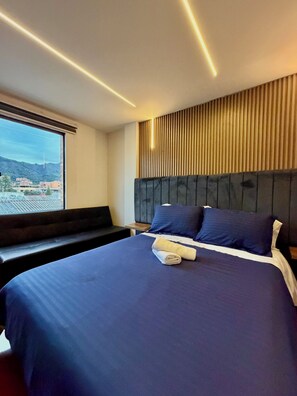 Une chambre d’hôtel moderne avec un grand lit, une tête de lit ornée de lignes verticales, une fenêtre donnant sur des immeubles et un banc placé près de la fenêtre.