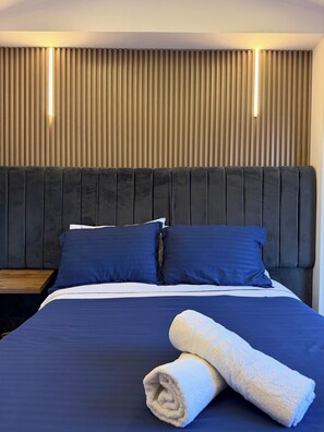 Una cama bien tendida con ropa de cama azul oscuro, dos almohadas y una toalla enrollada.