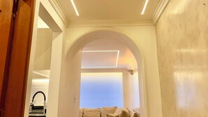 Un couloir avec une porte en bois, un canapé et une vue sur une pièce dont le plafond est blanc et qui est dotée d’un éclairage encastré.