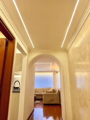 Un couloir avec une porte en bois, un canapé et une vue sur une pièce dont le plafond est blanc et qui est dotée d’un éclairage encastré.