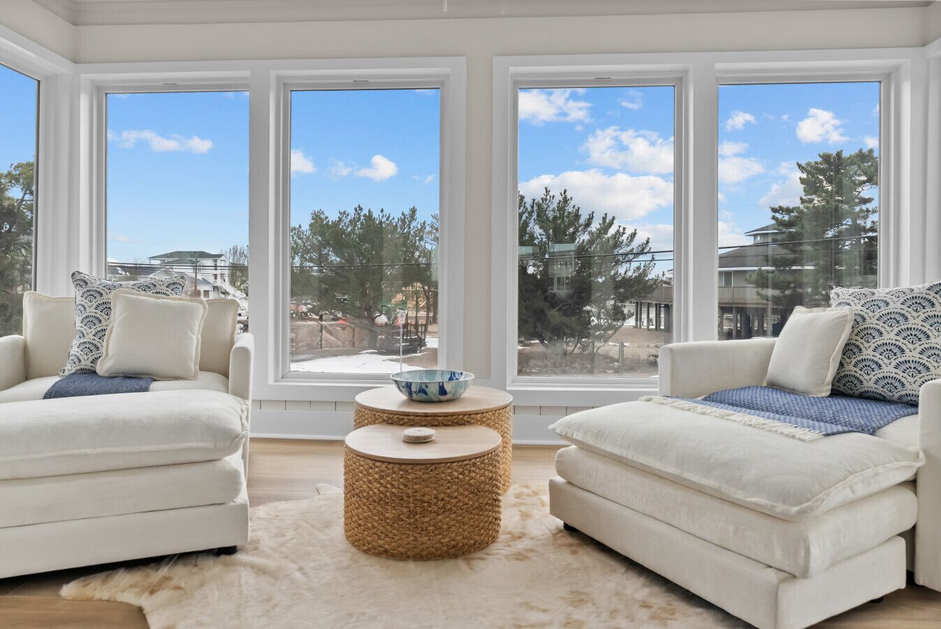 Un salón moderno con dos sofás blancos, una mesa de centro tejida y ventanas amplias que dan a vistas de árboles y edificios.