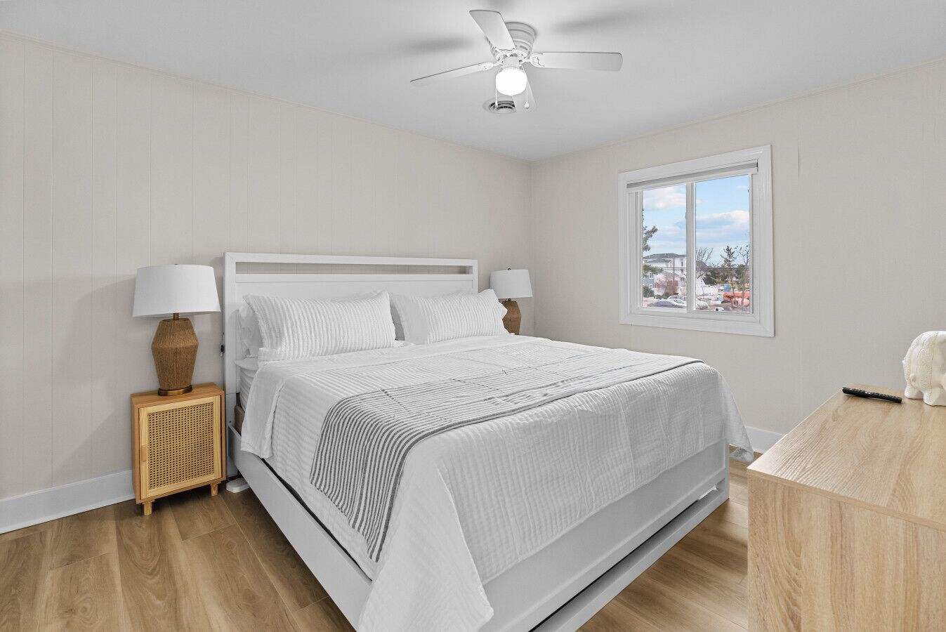 Un dormitorio con cama, lámparas de noche, ventilador de techo, ventana con vistas y una mesita con un control remoto.