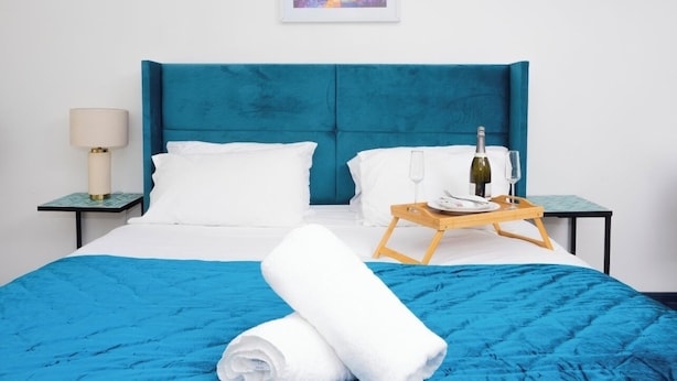 A neatly made bed with a blue headboard, white pillows, and a blue bedspread. There is a wooden tray with a bottle and glass on the bed. A framed picture hangs on the wall above the bed. - Ramsgate boutique  (Ramsgate)