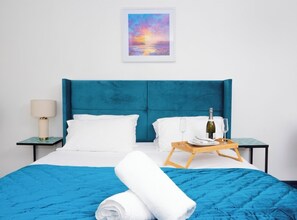 A neatly made bed with a blue headboard, white pillows, and a blue bedspread. There is a wooden tray with a bottle and glass on the bed. A framed picture hangs on the wall above the bed. - Ramsgate boutique (Ramsgate)