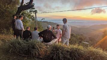Un groupe de personnes est assis et se tient debout sur une colline herbeuse, d’où l’on a une vue sur une vallée, avec un coucher de soleil en arrière-plan.