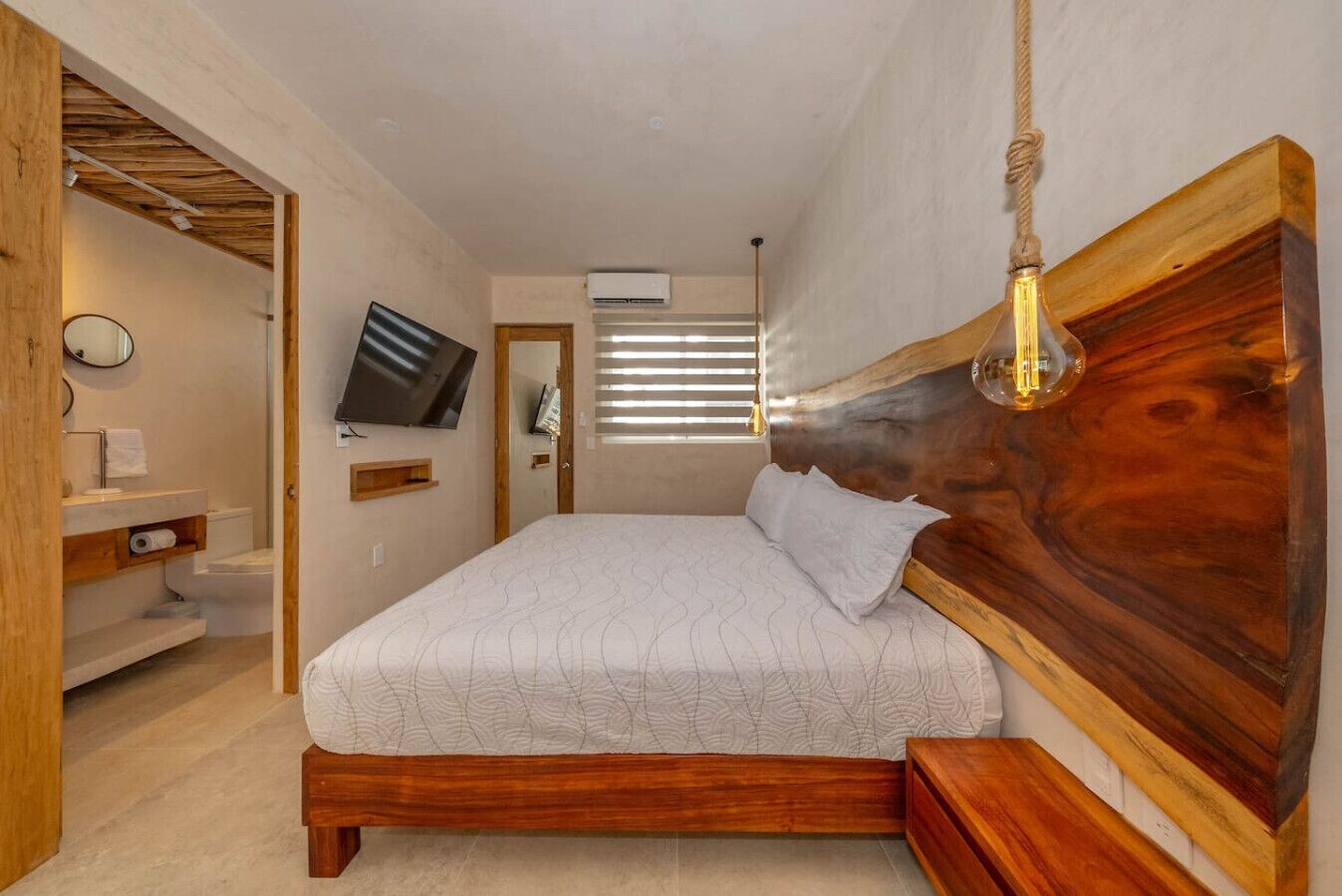 A bedroom with a wooden headboard, a bed with a patterned bedspread, a television mounted on the wall, a bathroom with a toilet and sink, and a window with blinds.