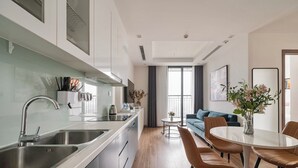 A modern kitchen with stainless steel appliances, a dining area with a round table and leather chairs, and a living room with a sofa and a large window. - Sunny - Vinhomes GreenBay Residences (Hanoi)