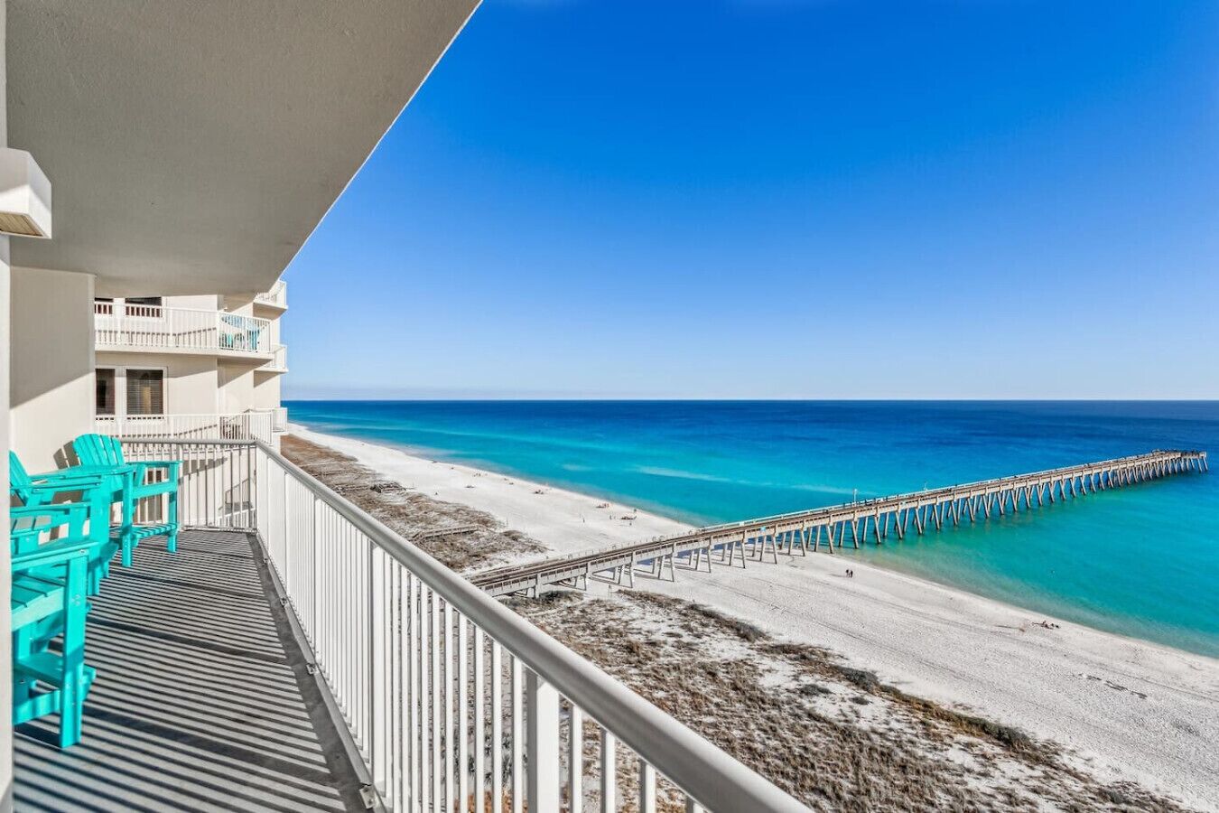A balcony with turquoise chairs overlooking a beach with a pier.