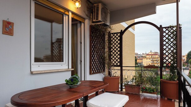 En balkon med et træbord og to hvide polstrede stole, en potteplante og udsigt til bygninger i baggrunden.