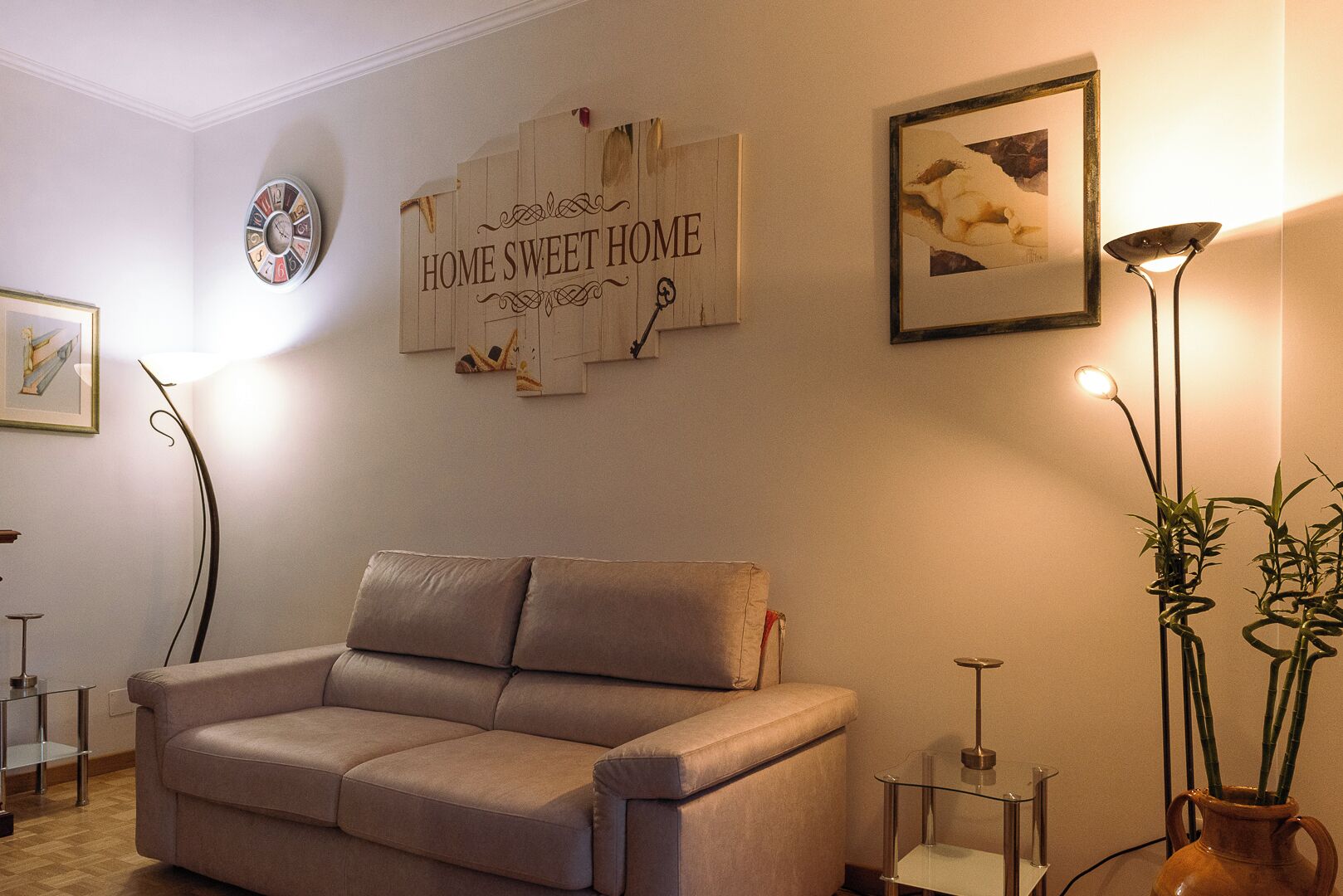 A living room with a beige sofa, a wall clock, a framed picture, a floor lamp, and a vase with plants.