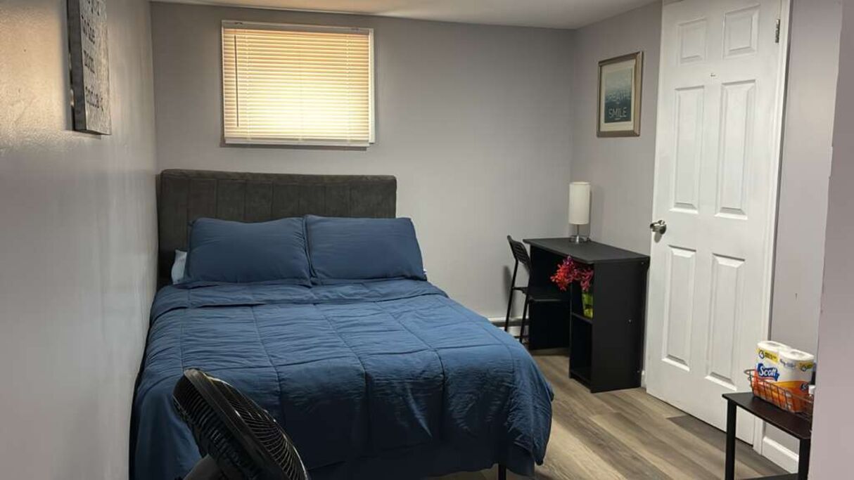 Une chambre à coucher comprenant un lit, un bureau, une chaise, un ventilateur, une fenêtre avec des stores et une porte.