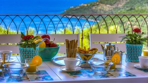 A breakfast table with fruits, pastries, and beverages, overlooking a scenic coastal view.