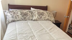 A bed with a white quilted mattress and floral-patterned pillows, a wooden headboard, a bedside table with a lamp, and a door to the right. - Charming 1-bedroom in a private house in Queens with AC, WiFi. Enjoy your stay (Queens)