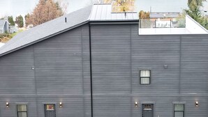 Un edificio grande y de color oscuro con techo plano, múltiples ventanas y vistas a un lago y colinas boscosas al fondo.