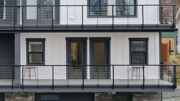 Un edificio residencial moderno de dos plantas con techo plano, ventanas amplias y un balcón con barandilla de metal.