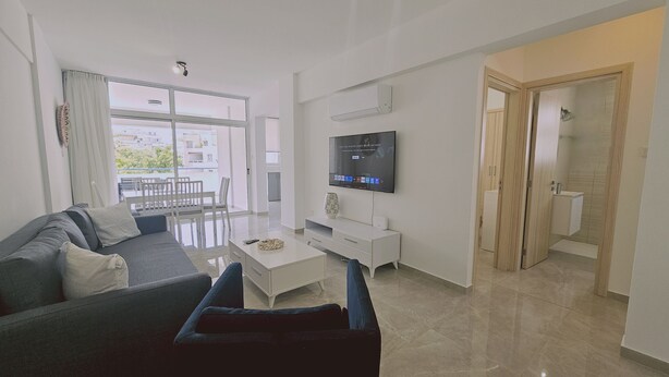 A modern living room with a sofa, coffee table, and TV. There is a balcony with a view of buildings and greenery. - 154 LCA - Stay Local Apartment (Larnaca)