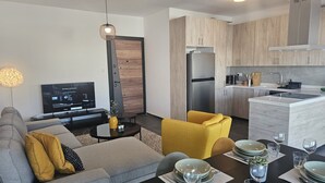 Ett modernt vardagsrum med en soffa, en gul fåtölj och ett matbord med plats för fyra personer. En platt-TV hänger på väggen, och ett kök med träskåp och vitvaror i rostfritt stål syns.