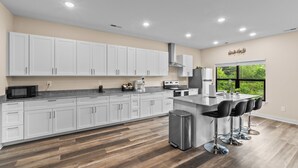 A modern kitchen with white cabinets, a central island with bar stools, stainless steel appliances, and a large window offering a view of greenery. - 3BR in Charlotte, 10 Min to Panthers Stadium Pet-Friendly (Charlotte)