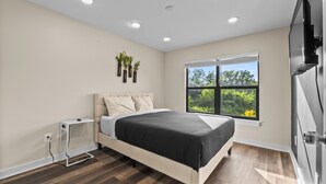 A modern bedroom with a bed, a TV, and a window with a view of greenery. - 3BR in Charlotte, 10 Min to Panthers Stadium Pet-Friendly (Charlotte)