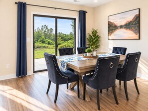 Un coin repas avec une table en bois, six chaises rembourrées et une vue sur la verdure grâce à de grandes portes vitrées.