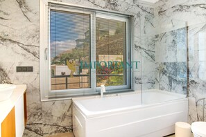 A bathroom with a marble wall, a bathtub, a window with blinds, and a toilet.