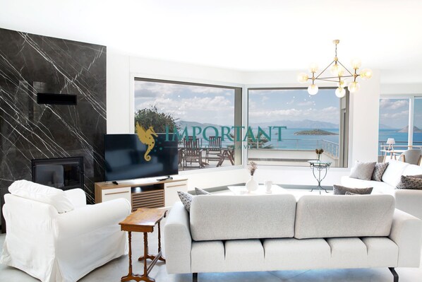 A modern living room with a large TV, comfortable seating, and a view of the sea.