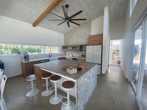 Una cocina moderna con una isla central, taburetes de bar, un ventilador de techo y vistas amplias a través de grandes ventanales.