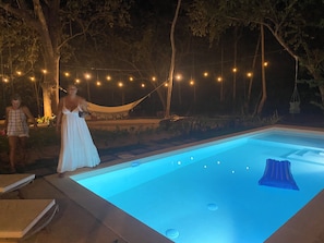 Una piscina con un tono azul, rodeada de árboles y luces colgantes.