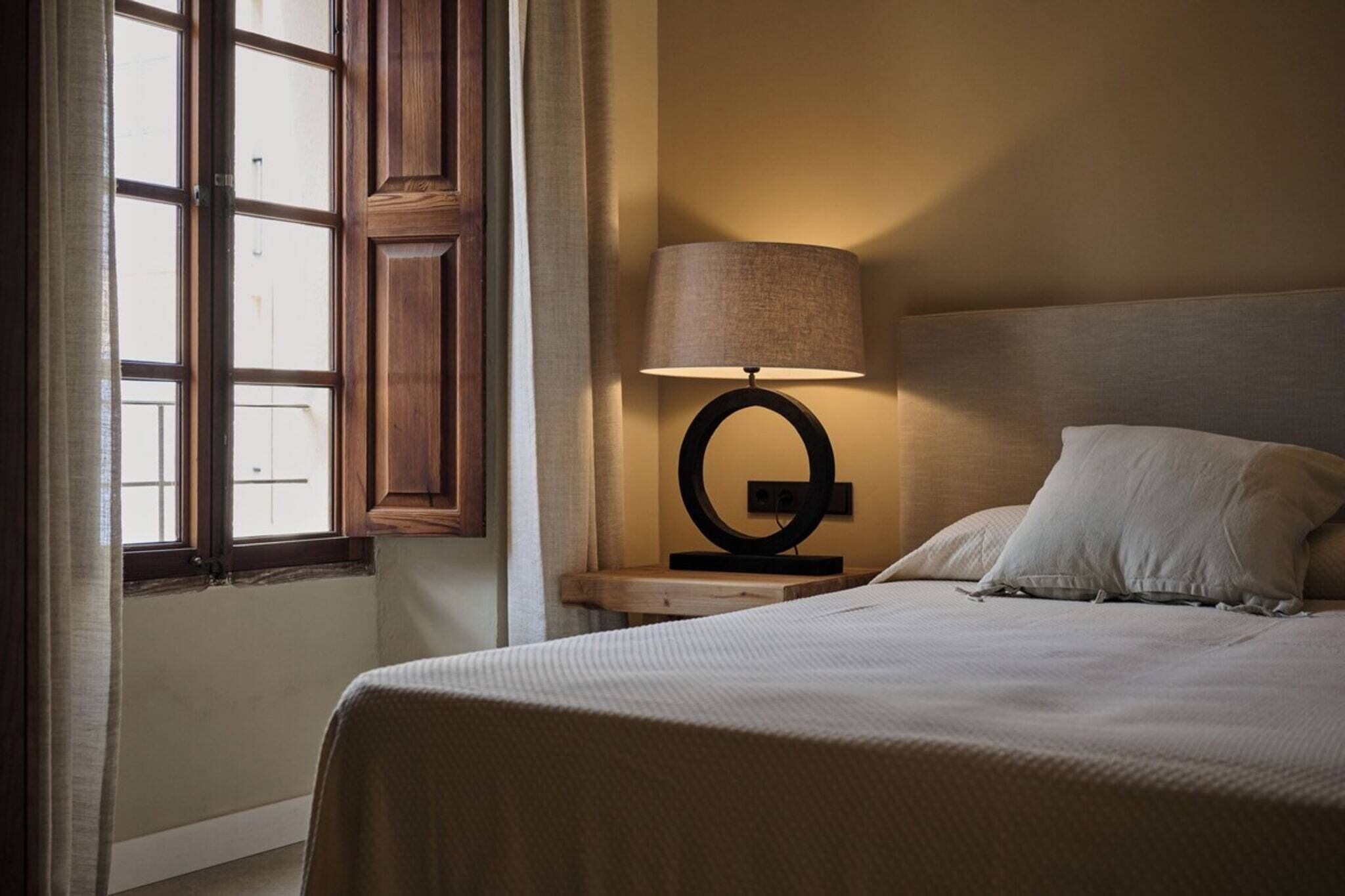 A hotel room with a bed, a nightstand with a lamp, a window with wooden shutters, and a view of the outside.