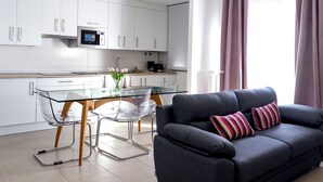 A modern kitchen with white cabinets, a glass dining table, and a grey sofa with striped cushions.
