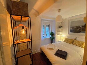 Ein Schlafzimmer mit einem Bett, einer Pendelleuchte, einem Fenster und einem an der Wand befestigten Regal mit einer Pflanze.