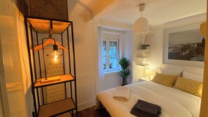 Une chambre à coucher avec un lit, un luminaire suspendu, une fenêtre et une étagère fixée au mur, sur laquelle se trouve une plante.