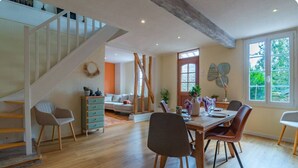 A spacious living area with a wooden staircase, a dining table with chairs, and a large window providing a view of the outdoors.