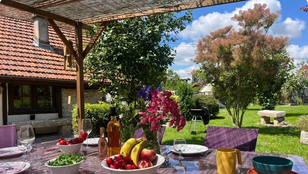 An outdoor dining table set with various dishes, a bottle, and a vase of flowers, under a pergola with a view of a garden and a house.