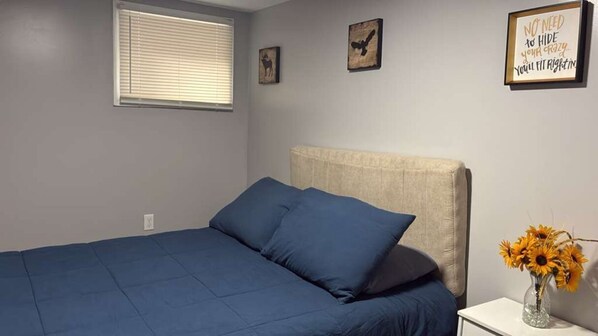 A bedroom with a bed, a nightstand, a window with blinds, and wall decorations. - Standard Room G | Near Prudential, EWR & NYC (Newark)